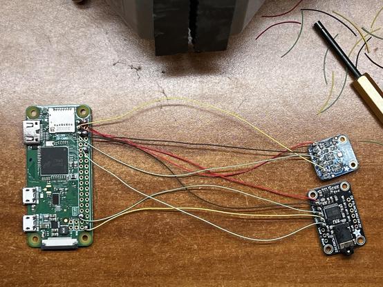 A raspberry pi zero W is wired to an audio DAC and a pressure/temperature/humidity sensor. Red, black, yellow, and green wire wrap wires are used to make the connections. The jaws of a vise, a manual wire wrap tool, and some stripped wire wrap wire insulation can be seen to one side.