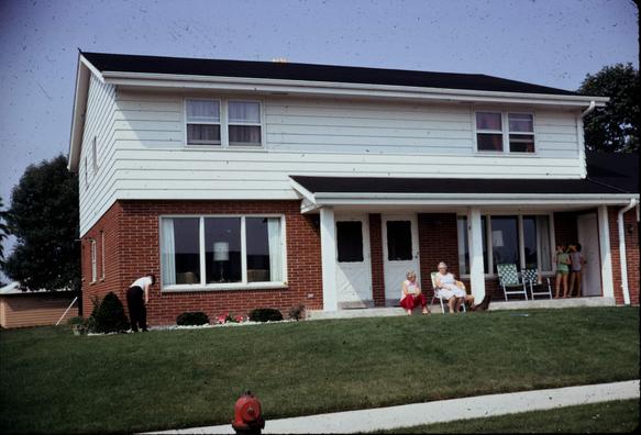 A 35mm color slide of a two story duplex. White siding on the top floor. Brick on the bottom store. Two white doors in the middle with house numbers. On the one to the right is a family gathered. A woman is styling an older woman's hair.