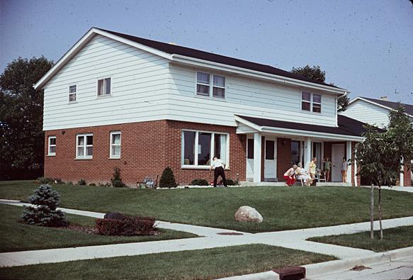 The duplex house again but this time with the curb visible. On the verge by the sidewalk is a sapling.