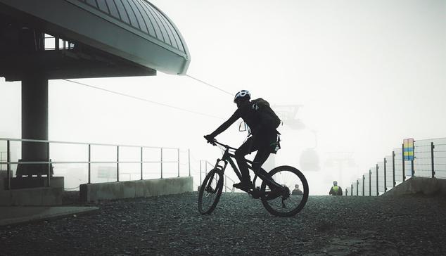 Silhouette of a mountain biker riding on gravel beside the top of a mountain lift, in front of a bright fog settling into the top of the mountain. A lift can be seen fading into the fog in the distance. The edge of the structure the lift chairs ride to can be seen on the left. The surrounding structures and gravel are dark and look almost metallic in color