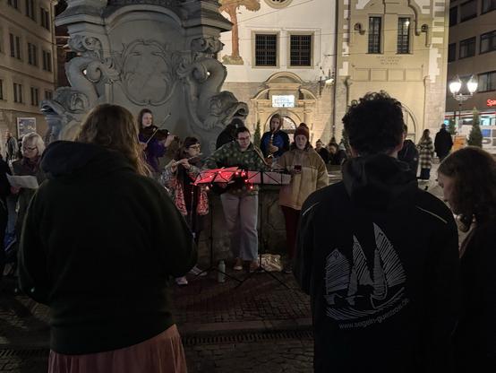 Vor dem Würzburger Vierröhrenbrunnen steht eine Gruppe junger Menschen mit Instrumenten und musiziert und singt zusammen mit den Menschen um sie herum.