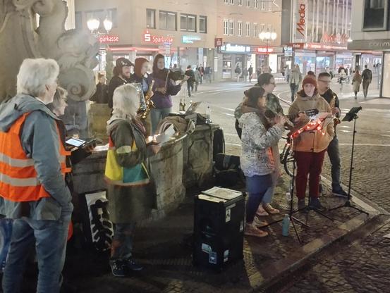 Mehrere Ordner:innen stehen am Brunnen vor dem Rathaus und singen mit.