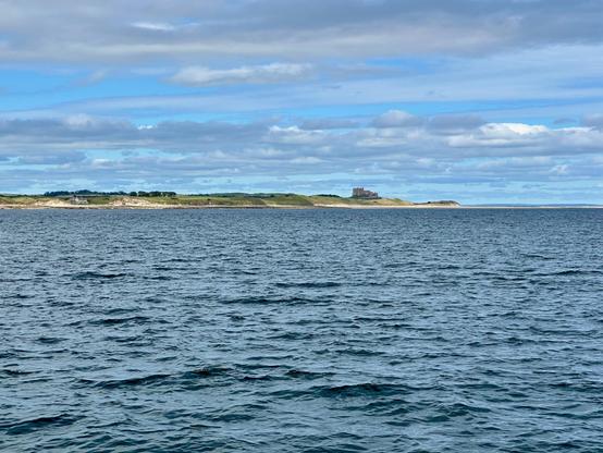 The image depicts a serene coastal scene with calm waters in the foreground and a distant shoreline. The horizon features a low-lying stretch of land with a faint outline of Bamburgh Castle visible towards the center. The sky above is partly cloudy, with patches of blue peeking through, creating a peaceful and expansive atmosphere.