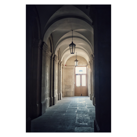 This is a portrait format colour image of a flagged stone corridor with vaulted stone ceiling. Ornate lamps hang from the stonework. At the end of the corridor are double half-glazed wooden doors which bring light into the composition. The architecture at the back of the image is highlighted by the daylight from the door, which also reflects off the smooth flagstones. The foreground is in shadow, creating a frame within a frame. Taken in Greenwich, London, with a Sony RX100.