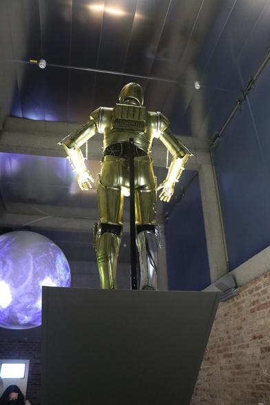 Der Droide C3PO steht auf einem Podest, wird von unten angestrahlt. Er wurde von hinten/unten fotografiert. Im Hintergrund sind eine projezierte Weltkugel sowie Teile der Architektur des Raumes zu sehen.