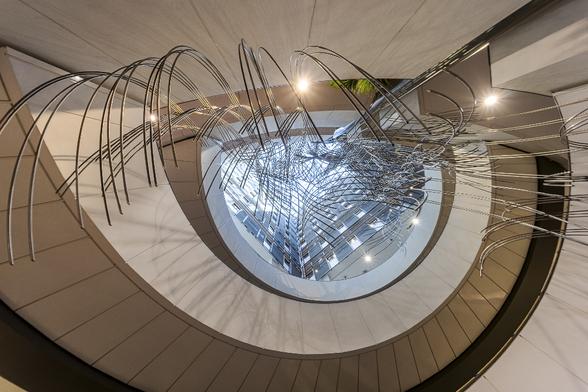 Look-up view on the central stairs in the European Parliament building in Brussels. The staircase has a semi-circle shape in soft warm tones that looks similar to an eye while the view on the upper floor in the middle looks slightly blueish and sparkling under a canopy  on a sunny day with a blue sky. A sculpture made of entangled metal line curves comes down from the ceiling to the bottom of the staircase, shapping an eyelash. This sculpture is meant to represent codecision and democracy.