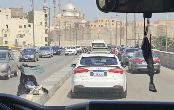 Photo taken from the backseat of a car during day. Between the rear-view mirror (equipped with trinkets and a tree-shaped air freshener) and the dashboard you can see a wide road filled with a traffic jam of an endless amount of cars. The middle of the road is marked by a prominent big stone step. Right next to the car ahead, a woman in wide black clothing sits on that step. She holds a small child on her lap and a broken green umbrella over her head to protect them from the sun. Full plastic bags sit in front of her. She seems unconcerned in the middle of the traffic. In the distance beyond the cars sand coloured limestone houses rise up and on top of a hill sits the impressive Alabaster Mosque or Muhammad Ali Mosque with its bright round domes and high minarets. Everything looks muted and dusty, even the blue sky above.