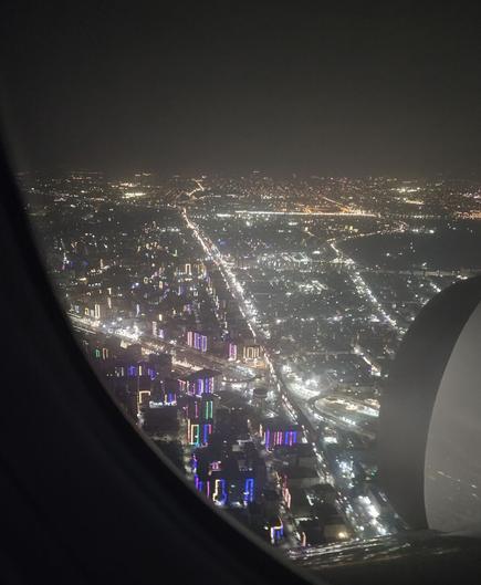 Photo out of the window of a plane by night. Berween the curved window frame and the round plane engine are the streets of Cairo visible. Two main roads are brightly illuminated by streetlights. One from top to bottom, one from roughly side to side. Between them endless lights and smaler roads are sprinkling light into the dark. On the right side of the picture the lights are white. In the distance near the horizon the lights are more orange. In the closer part at the edge of the window countless sky scrapers stand out in multicoloured purple, blue and green light, as if their whole structure has been eqipped with fairy lights.