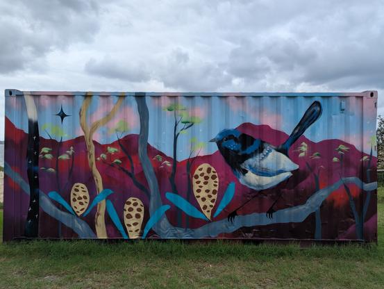 The image features a large, colorful mural painted on the side of a shipping container. The mural depicts a vibrant scene with a blue and black bird perched on a branch, surrounded by stylized trees and plants. The background is a gradient of blue and pink, with red hills and a starry sky. The bird is detailed with blue and black feathers, and the branch it sits on is painted in shades of blue and gray. The plants include large, spotted pods and green leaves, adding to the natural theme. The container is set against a grassy area with a cloudy sky above.