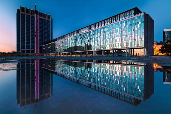 Reflection shot in a puddle of an administrative tower in Brussels and its rectangular annex, at the end of sunset when the sun still makes an orange glow on the windows, while the sky is turning dark turquoise. The building with its reflexion in the water evocates a giant spaceship from science fiction movies.