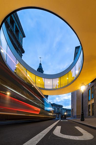 Low angle shot in slow motion of a bus passing through a circular illuminated passerelle stuck between two buildings in Brussels at blue hour. The yellow glow of the the passerelle makes a contrast with the darkening blue sky.