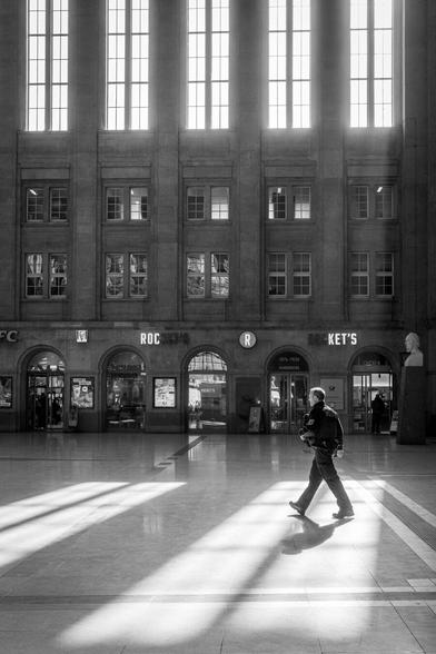 Menschen laufen durch die durch das Sonnenlicht durchflutete Bahnhofshalle des Leipziger Hauptbahnhofs. Eine Person steht direkt im Lichtkegel und wirft einen Schatten.