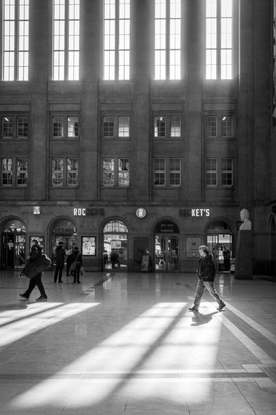 Menschen laufen durch die durch das Sonnenlicht durchflutete Bahnhofshalle des Leipziger Hauptbahnhofs. Eine Person steht direkt im Lichtkegel und wirft einen Schatten.