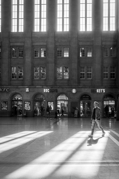 Menschen laufen durch die durch das Sonnenlicht durchflutete Bahnhofshalle des Leipziger Hauptbahnhofs. Eine Person steht direkt im Lichtkegel und wirft einen Schatten.