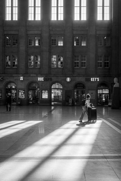 Menschen laufen durch die durch das Sonnenlicht durchflutete Bahnhofshalle des Leipziger Hauptbahnhofs. Eine Person steht direkt im Lichtkegel und wirft einen Schatten.