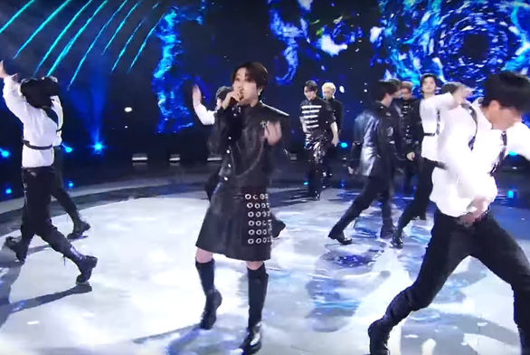 Han performing on stage during a music show, wearing an all-black outfit with black leather shorts that are cut to look like a short skirt