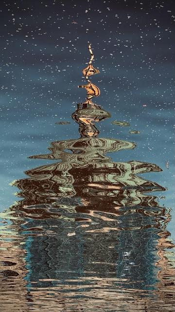 An artistic reflection of a tall structure in water, with swirling patterns and colors creating a mesmerizing effect. The surface appears disturbed, adding texture and depth to the scene.