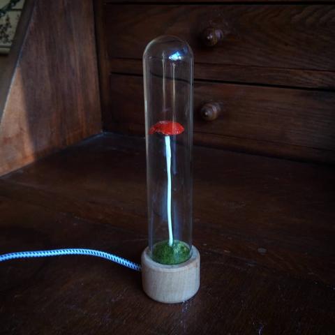 A small mushroom on a long white stalk sits under a tiny glass dome. At its base is a green mound from which it is growing, decorated with green stitches. A power cord trails to the left. The background is a dark wooden desk with drawers.