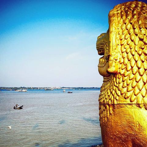 Photo shows the view from a golden guarding lion statue in Khmer art style, looking out on a water river view being the meeting water makes of the rivers Mekong and Tonle Sap. There are different kinds of river boats spread out in busy traffic. A single dove is visible in the sight direction flying by with a similar view. In the background the northern riverside is visible in a distance it Is a sunny day, with clear blue sky with some clouds in the horizon.