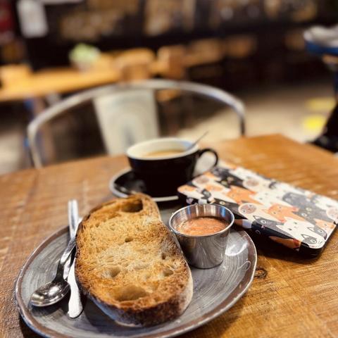Bodegón de desayuno con tostada en primer plano, con su aceite y tomate emulsionado, el café rico con leche fría de avena al fondo, y el Kobo en su funda de gaticos al lado.