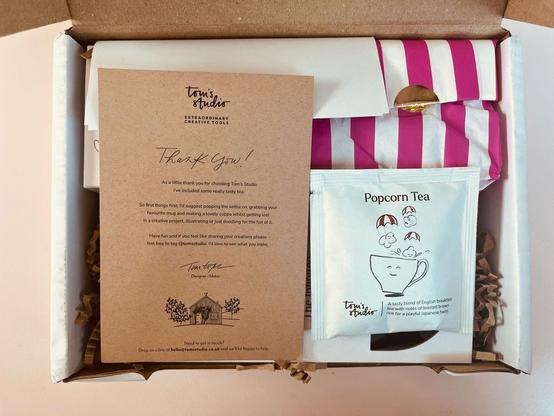 Blick von oben auf ein geöffnetes Paket. Obenauf liegen ein Beutel “Popcorn Tea” und eine Karte von Tom’s Studio, auf der die Aufforderung steht, sich erstmal in Ruhe eine Tasse Tee zu kochen und dann in einem kreativen Projekt zu versinken.