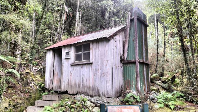 A Hatter 1 room cottage. Wooden walls. Corrugated iron chimney and roof. Sitting in the bush.