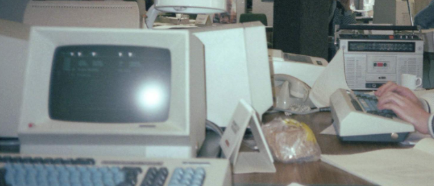 Detail van een werkplek in 1986 waarop links een dikke monochrome beeldscherm, dikke toetsenborden, een cassettespeler, printer en ouderwetse PTT telefoon te zien zijn. 

En een broodzak met boterhammen.