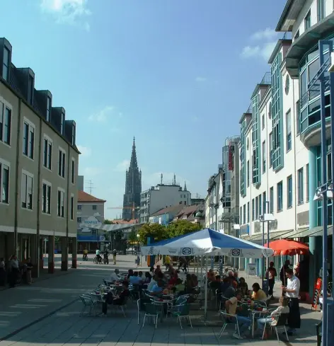 The image appears to depict a lively street or plaza scene in a town, characterized by modern buildings on both sides. There are outdoor seating areas with tables and umbrellas, where people are gathered, possibly enjoying food or drinks. In the background, there is a tall structure that could be a church or a tower, suggesting the presence of historical architecture nearby. The scene is bathed in sunlight, indicating it might be a pleasant day.

Image Credits: Wikimedia / Original uploader was Heinzkoch at de.wikipedia / CC BY-SA 3.0