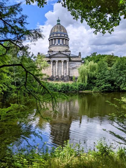 A medium, eye-level shot captures the serene Mausoleum in Dessau, Germany, reflected in the calm waters of a pond or small lake. The mausoleum, constructed from light-coloured stone, features a prominent dome at its apex and a classical facade with large columns. Its architectural style suggests a historical or memorial significance.
The surrounding environment is lush with greenery. Mature trees with vibrant green foliage frame the mausoleum on either side and in the foreground, with some branches partially obscuring the view, lending a natural and slightly wild feel to the scene. The water's surface mirrors the building and the sky above, creating a symmetrical and peaceful composition. The sky is partly cloudy, with patches of blue visible amongst the white clouds, suggesting a bright day. The overall atmosphere is one of tranquility and historical importance, nestled within a natural setting.