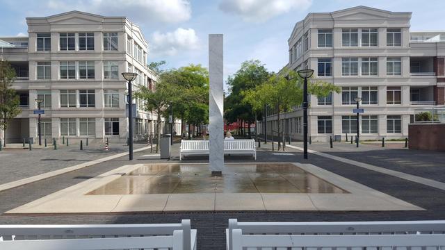 A photo of an empty square with a square marble tiled water feature with a high marble plinth in the middle of it. 

In the foreground the top of two sets of back to back benches lining the feature. On the opposite side a single set of similar benches in the middle, flanked by a black pole lamppost on the edge of the feature.

Sitting on the far bench facing away from the viewer, just visible to the right of the plinth, a person with light brown, shoulder lenght hair wearing a white jacquet. They are looking at a lane lined with trees between two four story, four floor, white stone buildings.