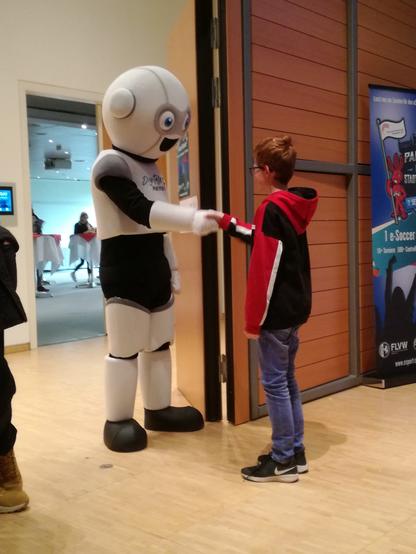 In einem Saal stehen sich ein zehnjähriger Junge mit blauer Jeans und schwarz-rotem Pulli und eine lebensgroße schwarz-weiße Roboter-Figur gegenüber und geben sich die Hand zur Begrüßung. Bei der Roboter-Figur handelt es sich um eine Werbeaktion des Heinz-Nixdorf-Museum-Forums, einem Computermuseum in Paderborn.