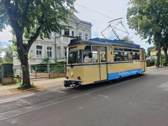 Eine einteilige alte Straßenbahn, die beige und blau lackiert ist. Im Hintergrund sind alte, ehrwürdige Einfamilien Häuser zu erkennen, sowie Bäume und ein bedeckter Himmel. Die Bahn ist in der Tat sehr alt und trägt die Ordnungsnummer 30. Die Zugzielanzeige ist sogar in Frakturschrift bedruckt. Außerdem ist die Liniennummer 87 sowie der Schriftzug "Woltersdorfer Straßenbahn" zu sehen. Das einzelne Gleis ist in Seitenlage in der Straße ein asphaltiert. Der Scherenstromabnehmer berührt die über dem Gleis hängende Oberleitung.