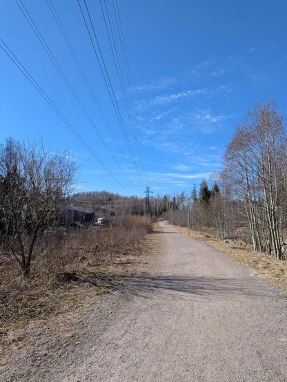 Asuinalueen vieressä kulkeva ulkoilureitti. Reitin vieressä kulkevat voimalinjat, jotka näkyvät mustina viivoina kirkkaan sinistä taivasta vasten.