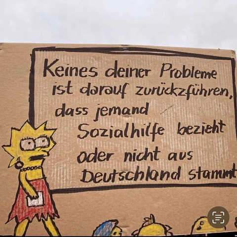 Pappkarton mit Lisa Simpson dozierend vor einem großen Schild oder Tafel: "Keines deiner Probleme ist darauf zurückzuführen, dass jemand Sozialhilfe bezieht oder nicht aus Deutschland stammt".