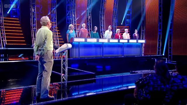 A long desk has seven people behind it; at the left of shot is a contestant at a podium, looking at right angles to the desk.