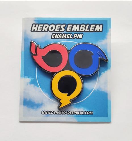 A photograph of an enamel pin depicting a stylized rounded version of the team emblem from Sonic Heroes. It's attached to a backing card that shows a blue sky with clouds, with the test "HEROES EMBLEM Enamel Pin" at the top, and "WWW.DYNAMO-DEEPBLUE.COM" at the bottom.