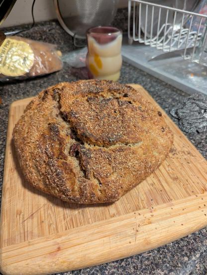 Bread 15B sitting on a cutting board, little wheat germ husks covering it with an appetizing split running along the sides. A pale ochre silver an pink gold glass cup in the background