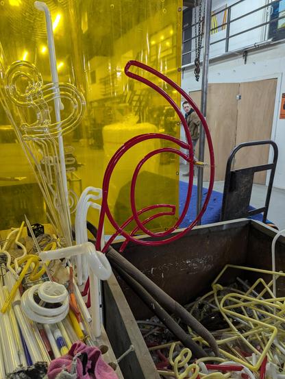 An upside down red neon "9" sitting in the cooling rack above the smashed reject bin on my bending table. Yellow welding screen in the background.
