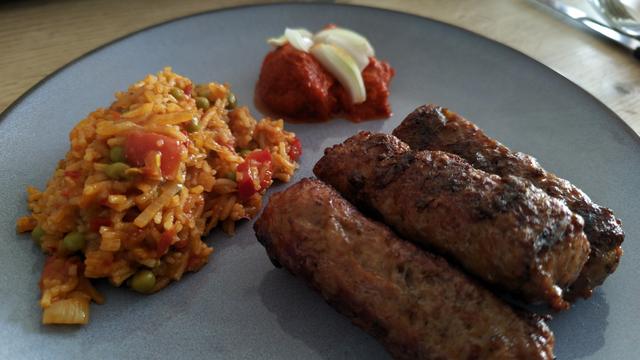 auf einem blauen Teller liegen 3 Cevapcici, etwas Duvicreis und Ajvar mit Zwiebeln