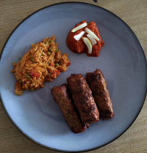 auf einem blauen Teller liegen 3 Cevapcici, etwas Duvicreis und Ajvar mit Zwiebeln