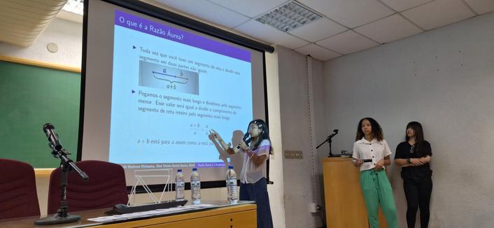 A foto mostra um auditório com telão projetando uma apresentação sobre o conceito de Razão Áurea. A apresentação inclui um diagrama e texto em português, com títulos e sub-títulos explicando o tema. No centro da imagem, uma adolescente está apontando para o diagrama no telão, enquanto fala. Ela está vestida com uma camiseta branca e calça jeans. À direita, duas outras adolescentes observam a apresentação, uma delas segurando um microfone.