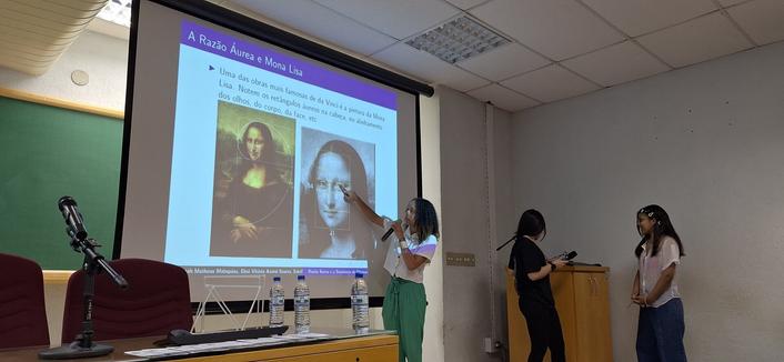 A foto mostra uma apresentação em um auditório. No centro da imagem, uma pessoa está apontando para uma projeção na tela, que exibe a obra "A Mona Lisa" de Leonardo da Vinci, com um título em português: "A Razão Áurea e a Mona Lisa". A projeção inclui uma explicação sobre a relação da Mona Lisa com a Razão Áurea, destacando retângulos e linhas nos olhos, corpo e cabelos da figura. A pessoa apontando está vestida com uma camiseta branca e calças verdes, segurando um marcador. À direita, duas outras pessoas estão de pé, uma delas usando um microfone, sugerindo que estão participando da apresentação.