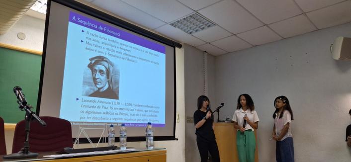 A foto mostra um auditório com um telão onde há uma apresentação sobre a "Sequência de Fibonacci". A tela exibe um slide com o título "A Sequência de Fibonacci" e uma imagem de Leonardo Fibonacci. O slide menciona a importância da sequência na arte e arquitetura, além de destacar a contribuição de Leonardo Fibonacci para a matemática. Na frente da tela, há três jovens, todas segurando microfones.