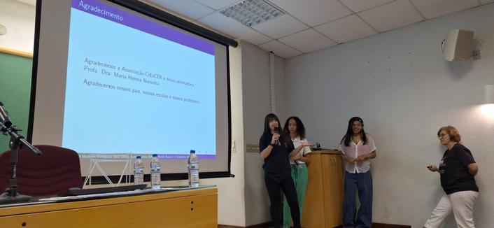 A foto mostra um auditório, com um grande telão projetando uma slide de agradecimento. O slide, contém o título "Agradecimento". Abaixo, está escrito: "Agradecemos à Associação CrEsCER e nossos orientadores Profª. Dra. Maria Helena Noronha. Agradecemos nossos pais, nossas escolas e nossos professores." A sala tem paredes brancas e um teto com iluminação fluorescente. No primeiro plano, há uma mesa com um microfone, uma cadeira vermelha e várias garrafas de água. Três jovens estão em pé, duas delas segurando microfones, e uma delas está apontando para o slide. Uma mulher mais velha está ao lado, observando.