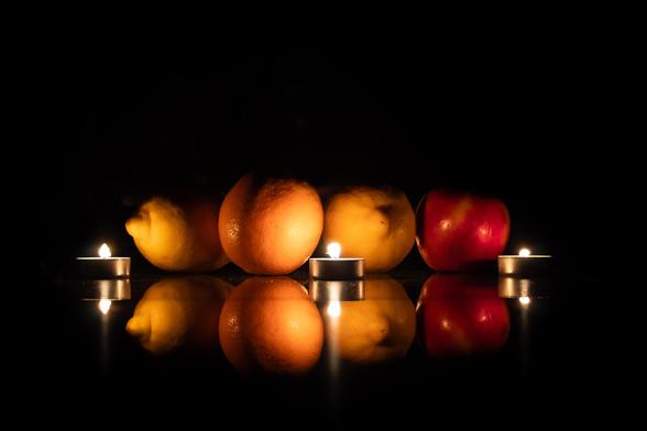 A series of fruits (two lemons, an orange and an apple) are seen under the light of candles. They are reflected in the surface they rest on.