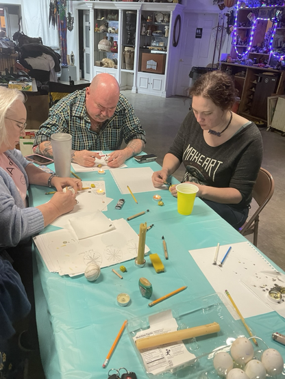 Two women and a man writing pysanky eggs. With a blue table cloth with eggs, paper, pencils, candle and kitskas