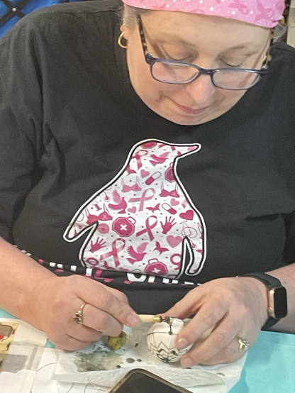 A woman wearing a gray shirt with a pink and white design and a pink bandanna writing pysanky egg. With a blue table cloth with eggs, paper, wax and kitskas