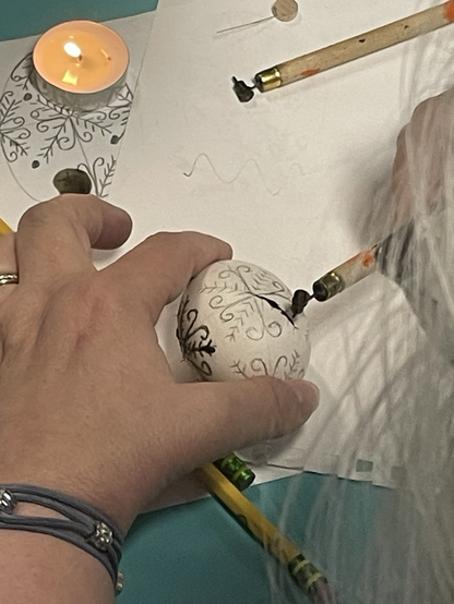 A woman writing her pysanka with a kitska. There are pencils on the table, a lit candle and she is wearing a bracelet