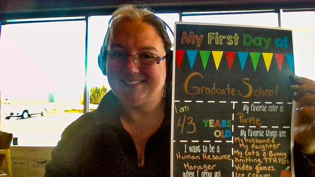 A woman with glasses and headphones smiles while holding a colorful sign that reads, "My First Day of Graduate School." The sign includes details such as her age (43), favorite color (purple), and favorite things. The background features large windows with natural light.