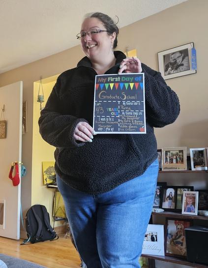 A woman with glasses and a black fleece jacket stands in a living room, smiling while holding a "My First Day of Graduate School" sign. The room has framed photos on a shelf, a backpack on the floor, and a partially open door in the background.